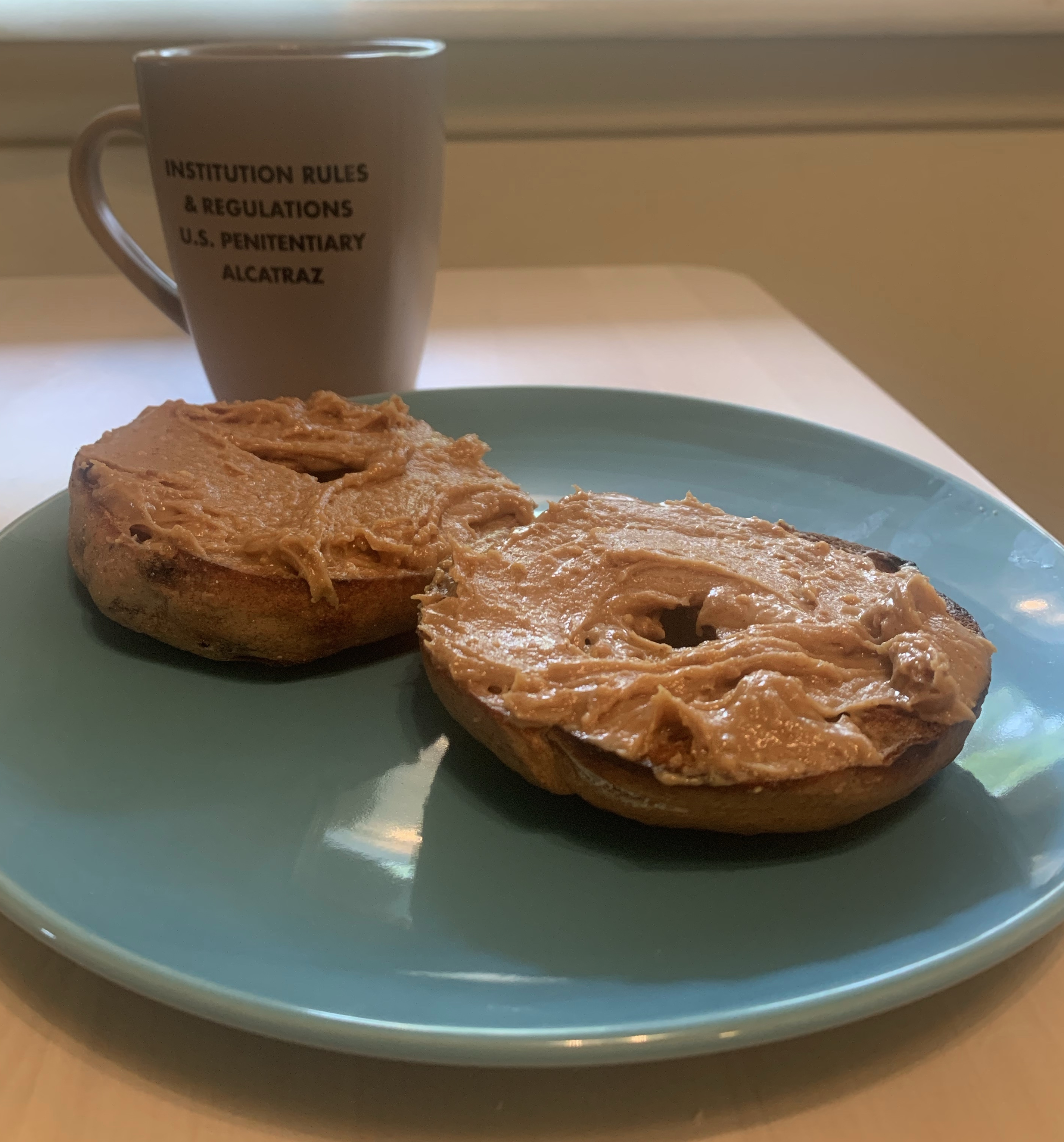 open faced bagel topped with peanut butter and honey on a blue plate in front of a coffee cup that reads "institution rules & regulations u.s. penitentiary alcatraz"