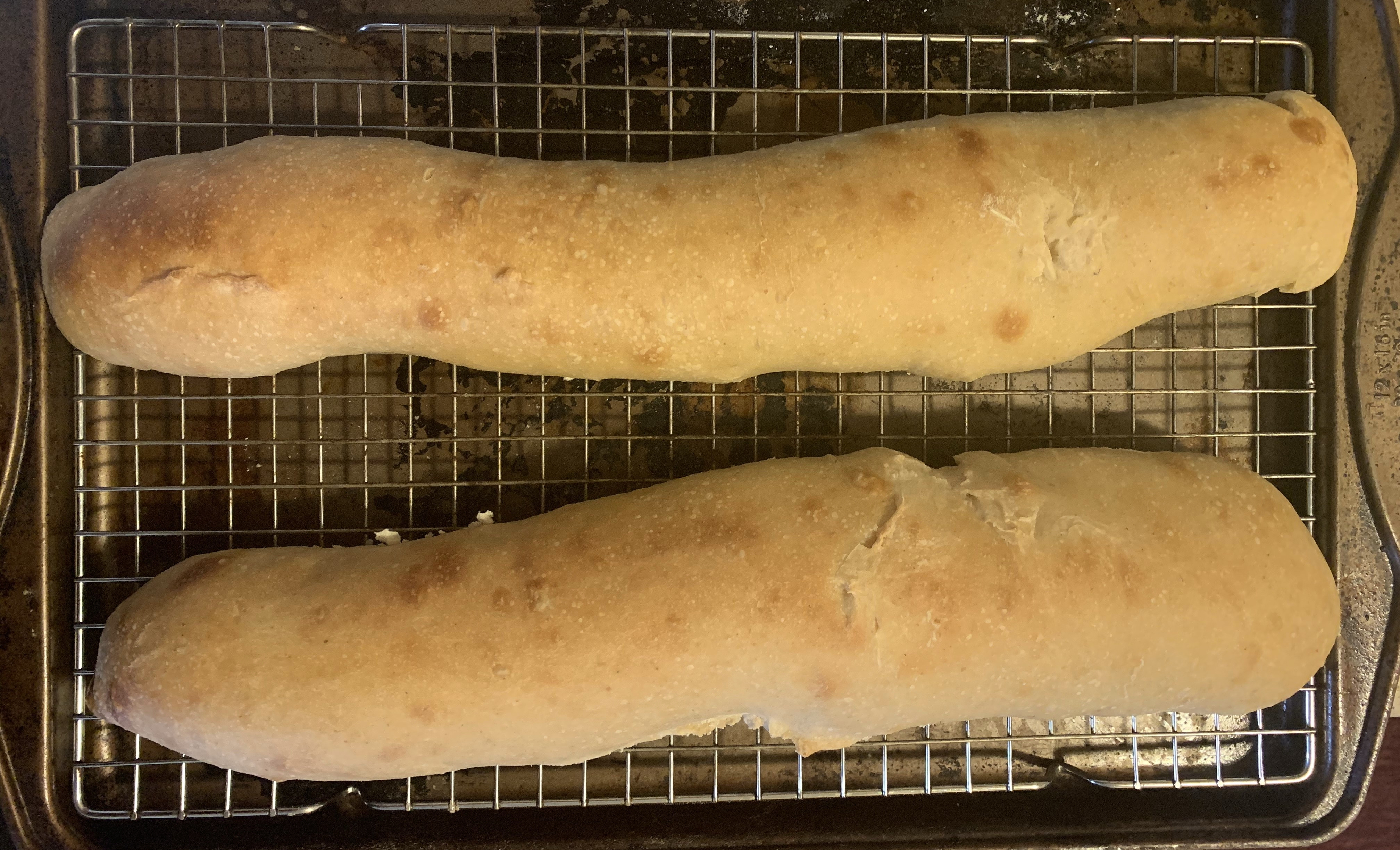 Two loaves of bread with golden brown crusts, sitting on a wire rack in a baking sheet.