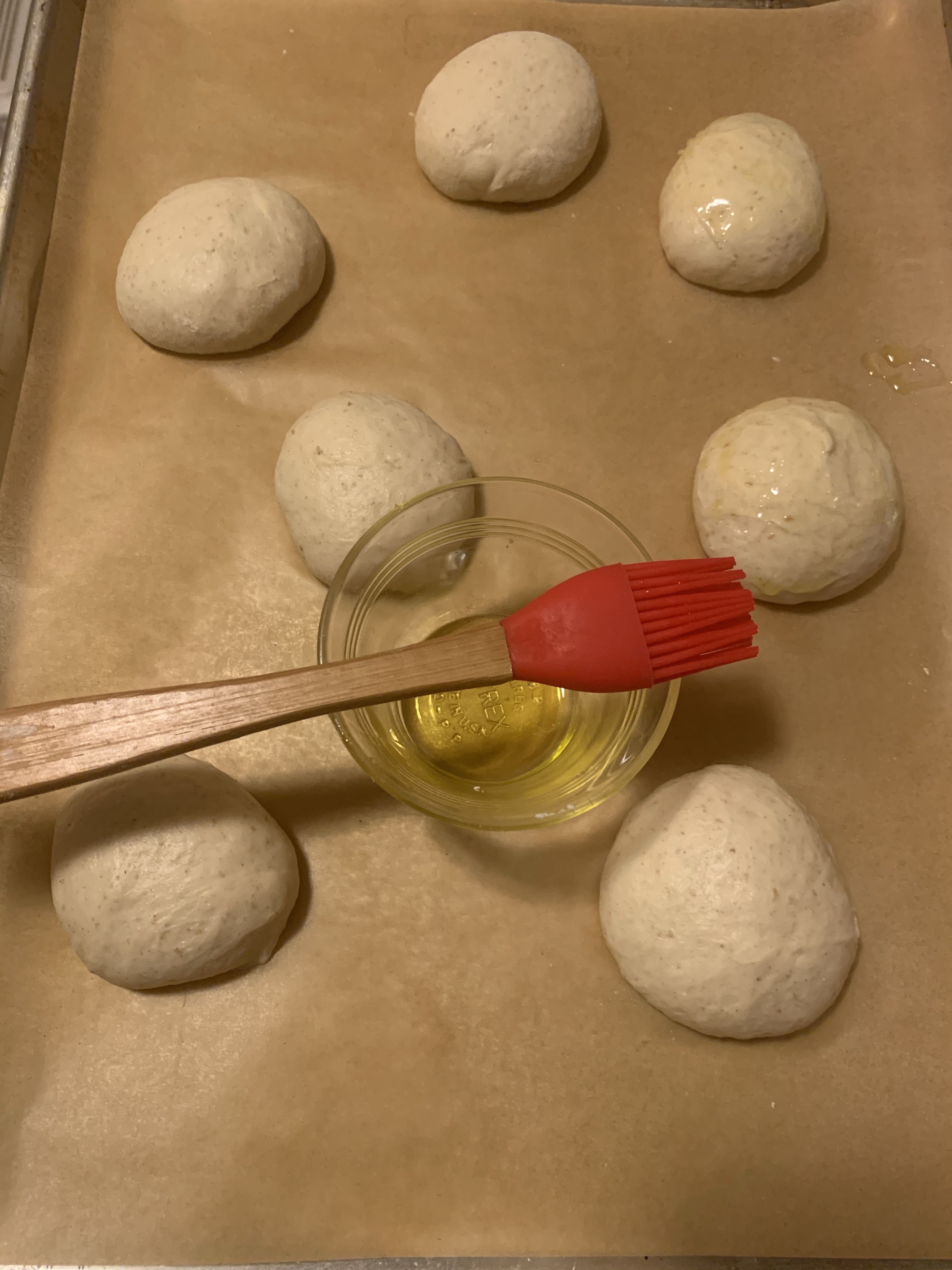 Seven rolls on parchment paper, some covered in oil. A glass dish of olive oil and rubber pastry brush are among the rolls.
